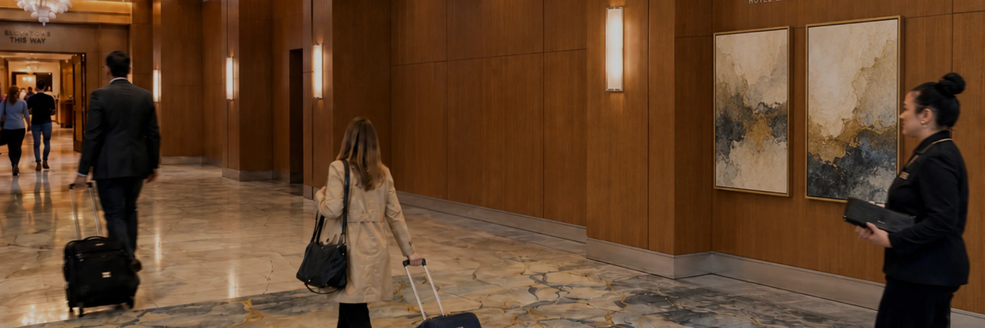 Hotel lobby corridor with wood-paneled walls; a woman in a beige trench pulls a rolling suitcase and carries a black bag, a man in front also with luggage, and a staff member stands by framed art on the right.