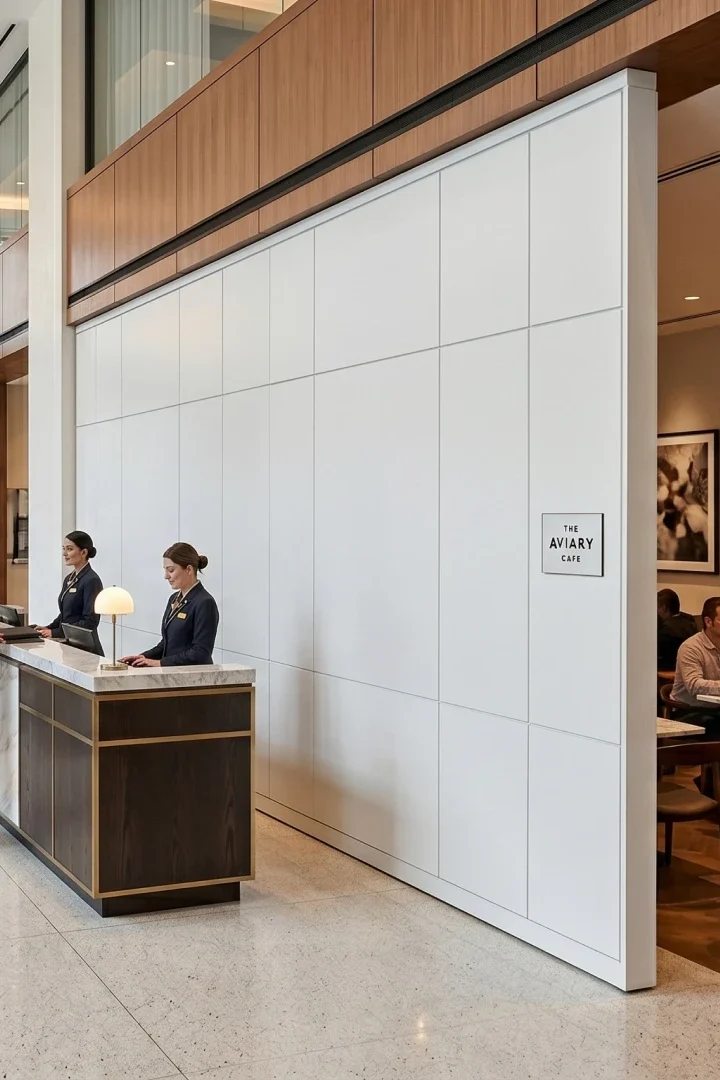 Modern hotel lobby reception: couple checks in at a marble-front desk, assisted by two attendants, white paneled wall behind.