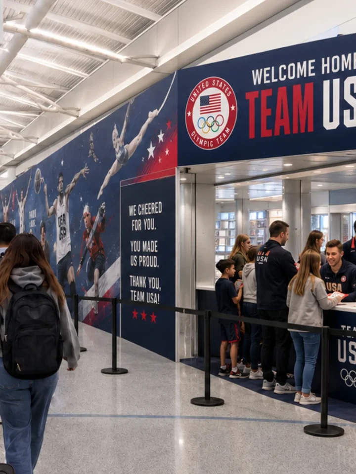 Airport arrivals area with a large 'Welcome Home Team USA' banner and Olympic logo, where travelers queue at a help desk.