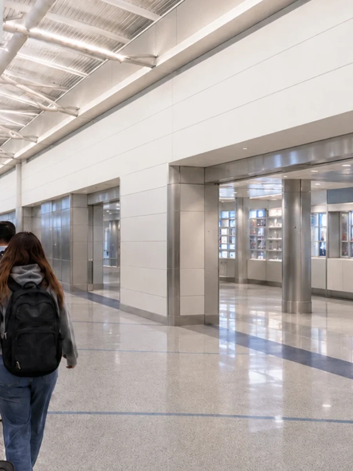 Travellers with rolling suitcases walk through a bright airport concourse with white walls and storefronts on the right.